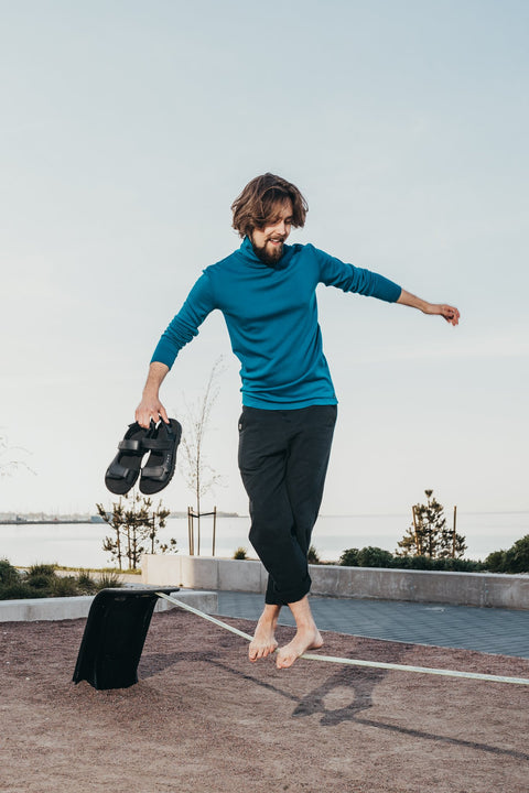 1. Man balancing on a beam holding Omaking Lauri Leather Sandals in black, showcasing sporty style and comfort