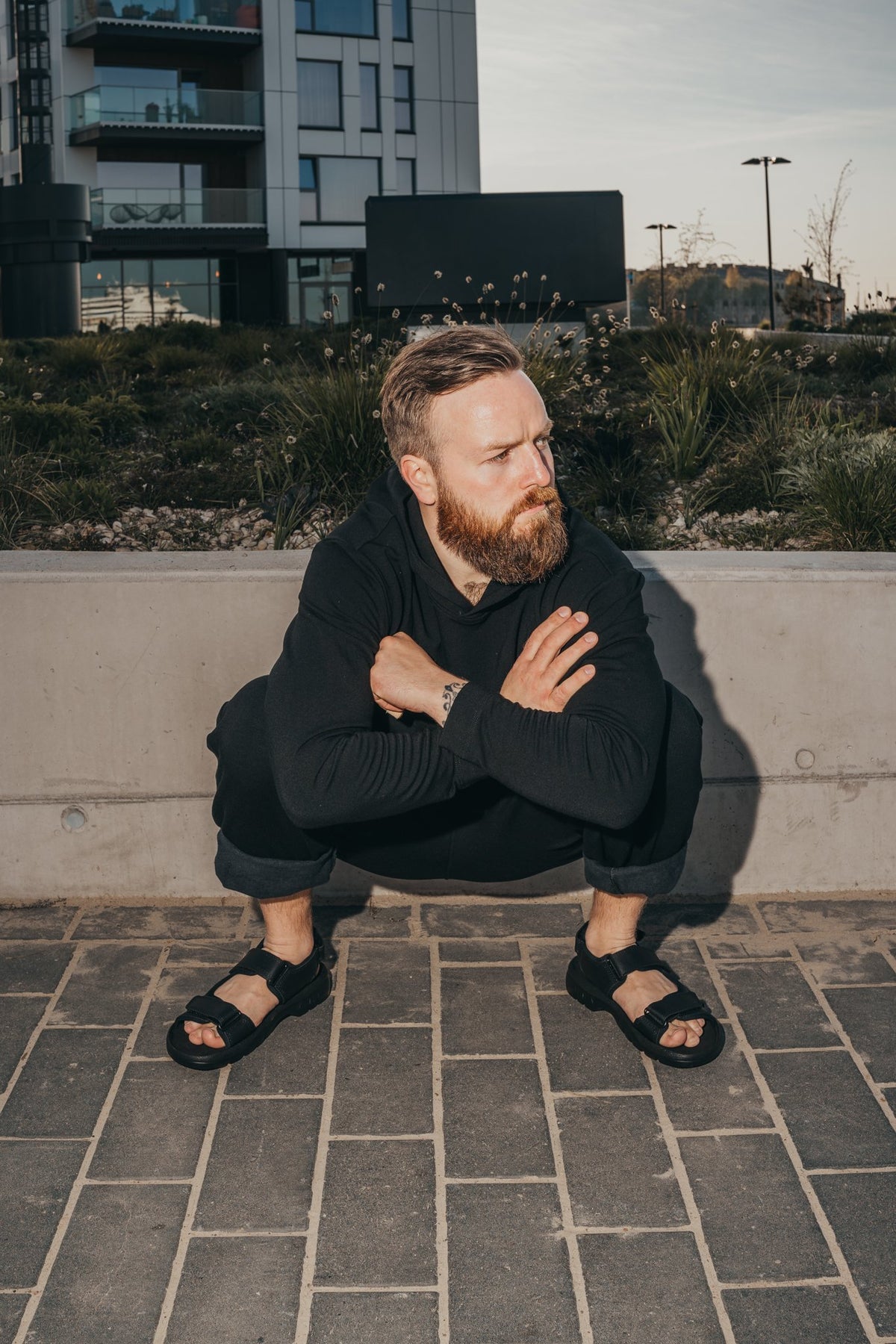 2. Man squatting outdoors wearing Omaking Lauri Leather Sandals in black, highlighting adjustable velcro straps