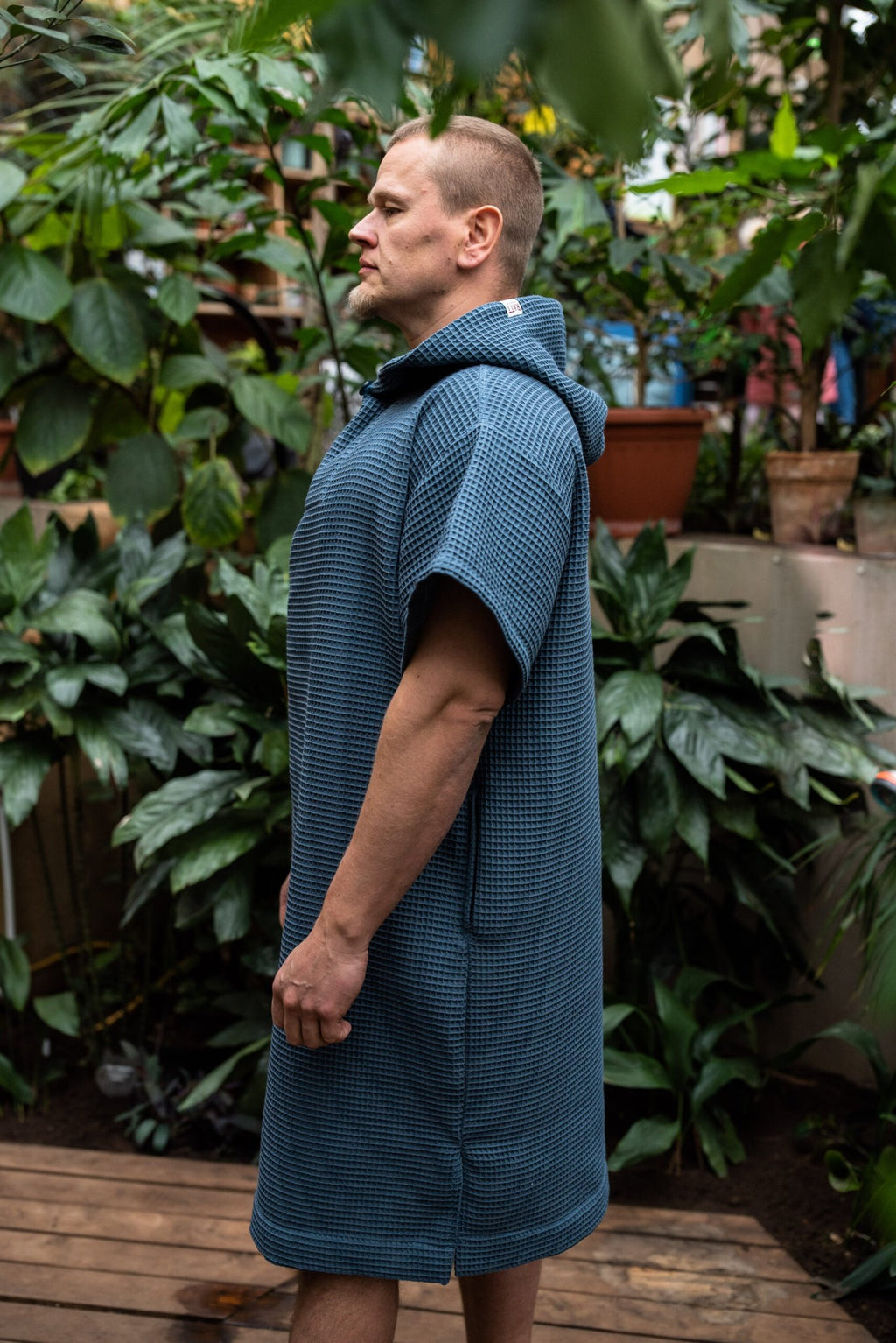 16. Man in blue RÄTT short-sleeve surf poncho with hood, side view in garden setting