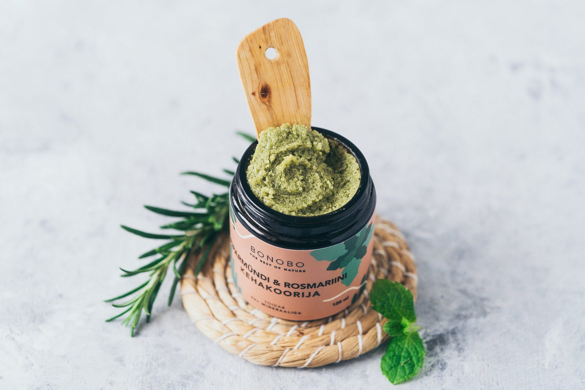 2. Open jar of BONOBO peppermint and rosemary body scrub with wooden spoon, surrounded by rosemary and mint
