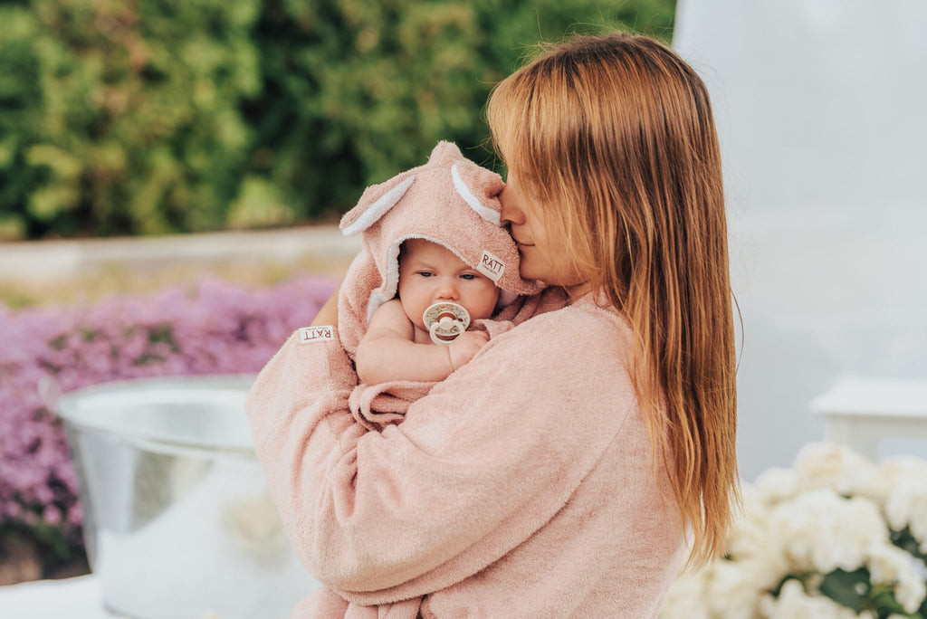 1. Mother holding baby wrapped in RÄTT hooded baby bath towel with rabbit ears, outdoor setting