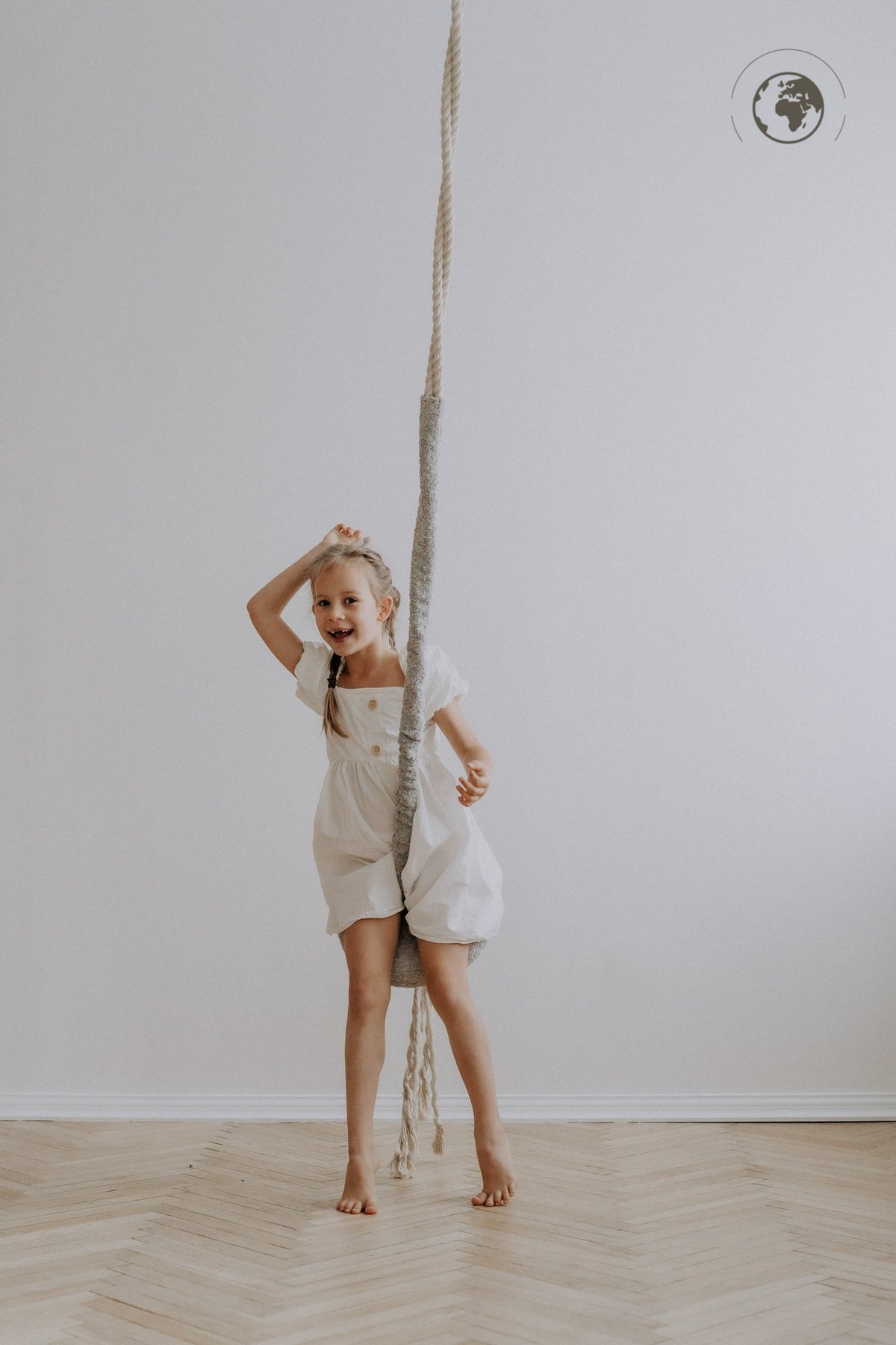 1. Girl in white dress standing with Nofi KIDS HopOn Swing in minimalist room, smiling and holding rope