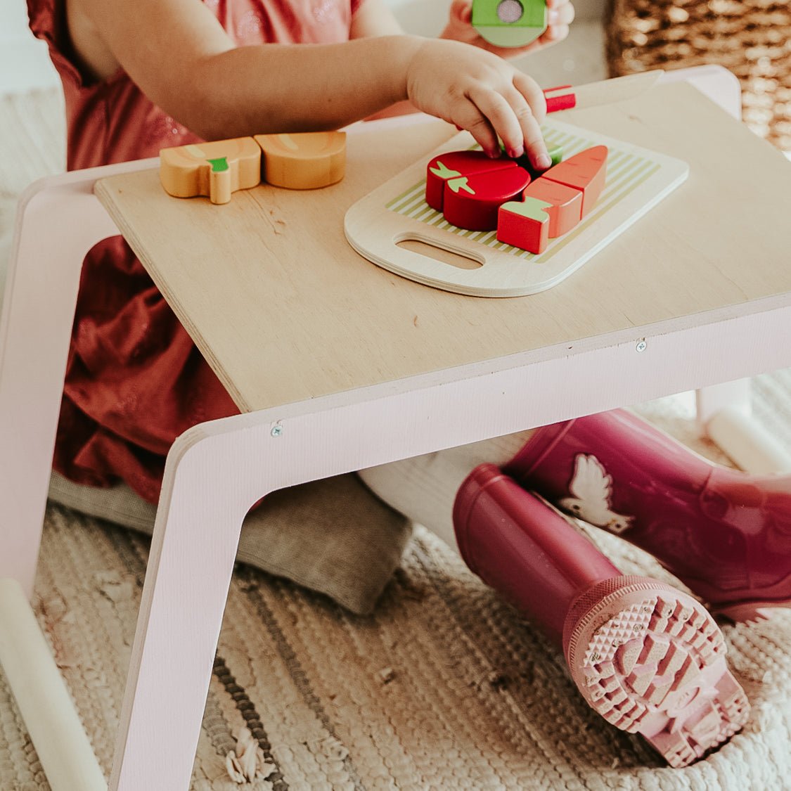 1. Child in pink dress and boots playing with toy food on small wooden desk