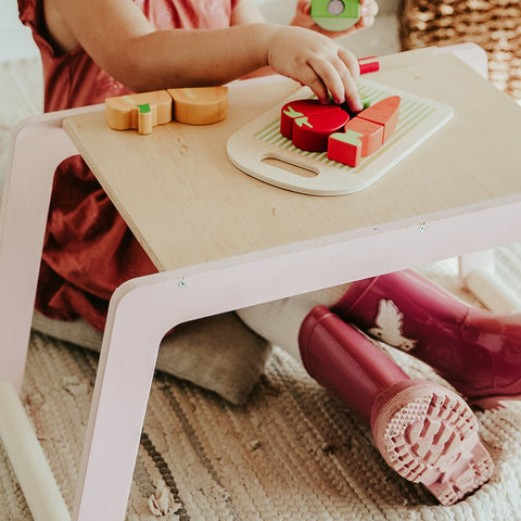 1. Child in pink dress and boots playing with toy food on small wooden desk