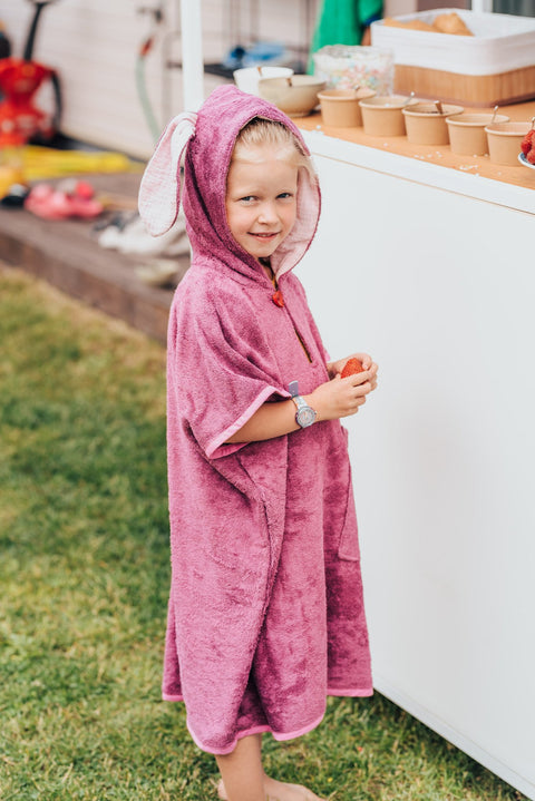 5. Child wearing pink RÄTT short-sleeve poncho with hood, standing outdoors