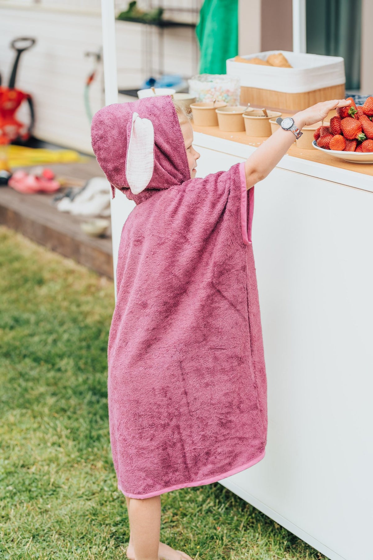 6. Back view of child in pink RÄTT short-sleeve poncho reaching for strawberries