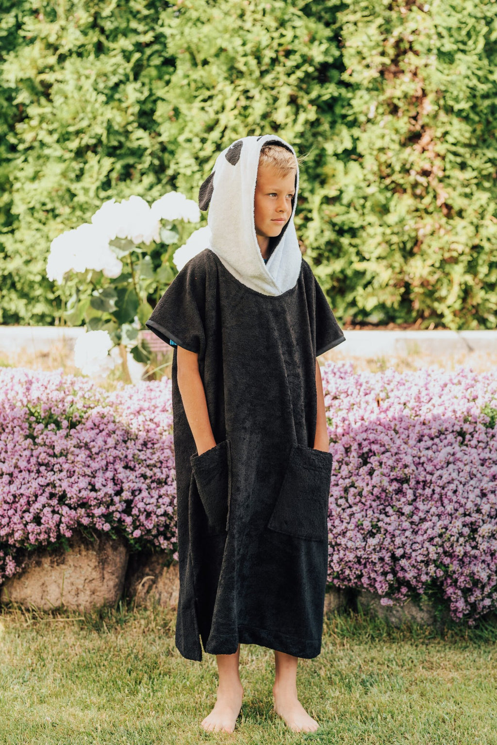 3. Child wearing RÄTT Short-Sleeve Poncho PANDA, standing in garden with hood up