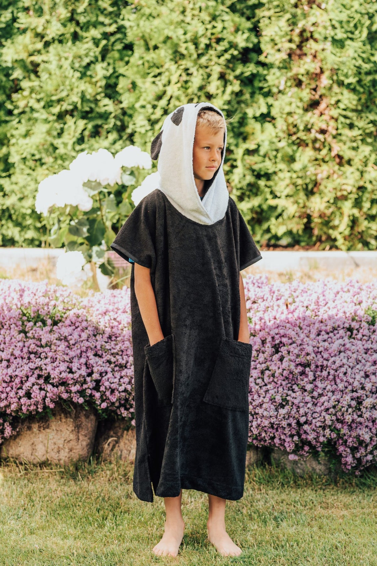 3. Child wearing RÄTT Short-Sleeve Poncho PANDA, standing in garden with hood up