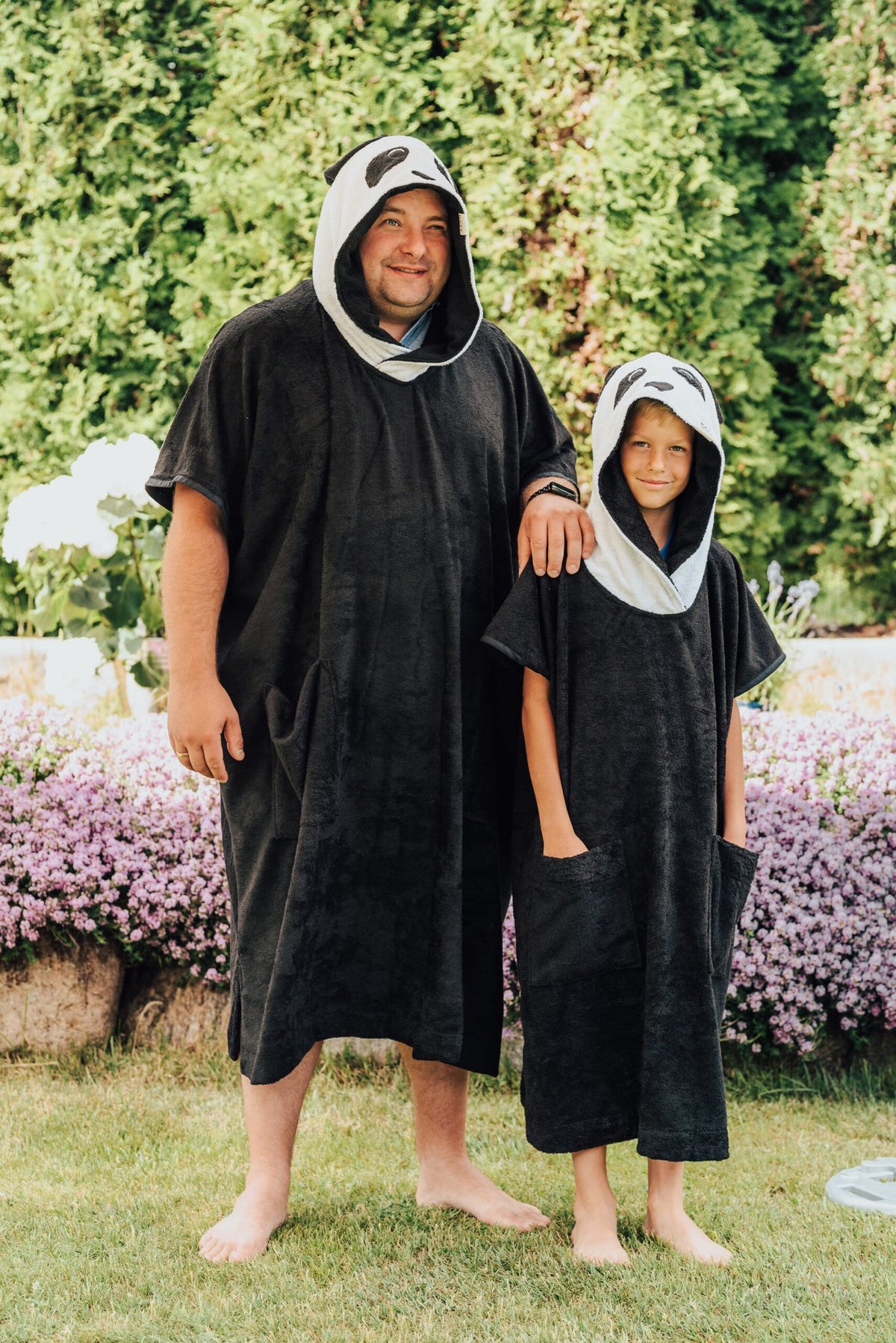2. Adult and child wearing RÄTT Short-Sleeve Poncho PANDA, standing back-to-back in garden