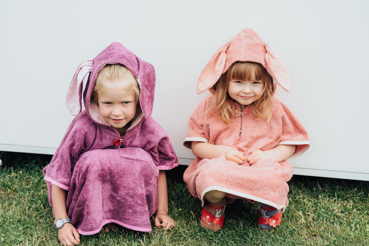 7. Two children in RÄTT short-sleeve ponchos, pink and purple, sitting on grass
