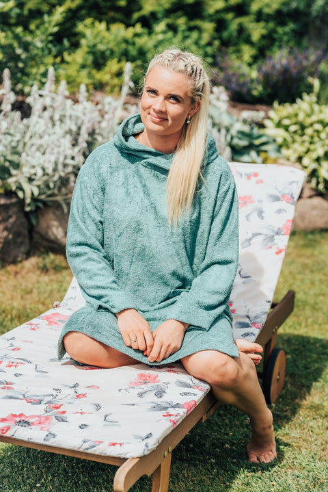 2. Women's bamboo terry long-sleeve poncho in green, worn by model sitting on a floral lounge chair in garden
