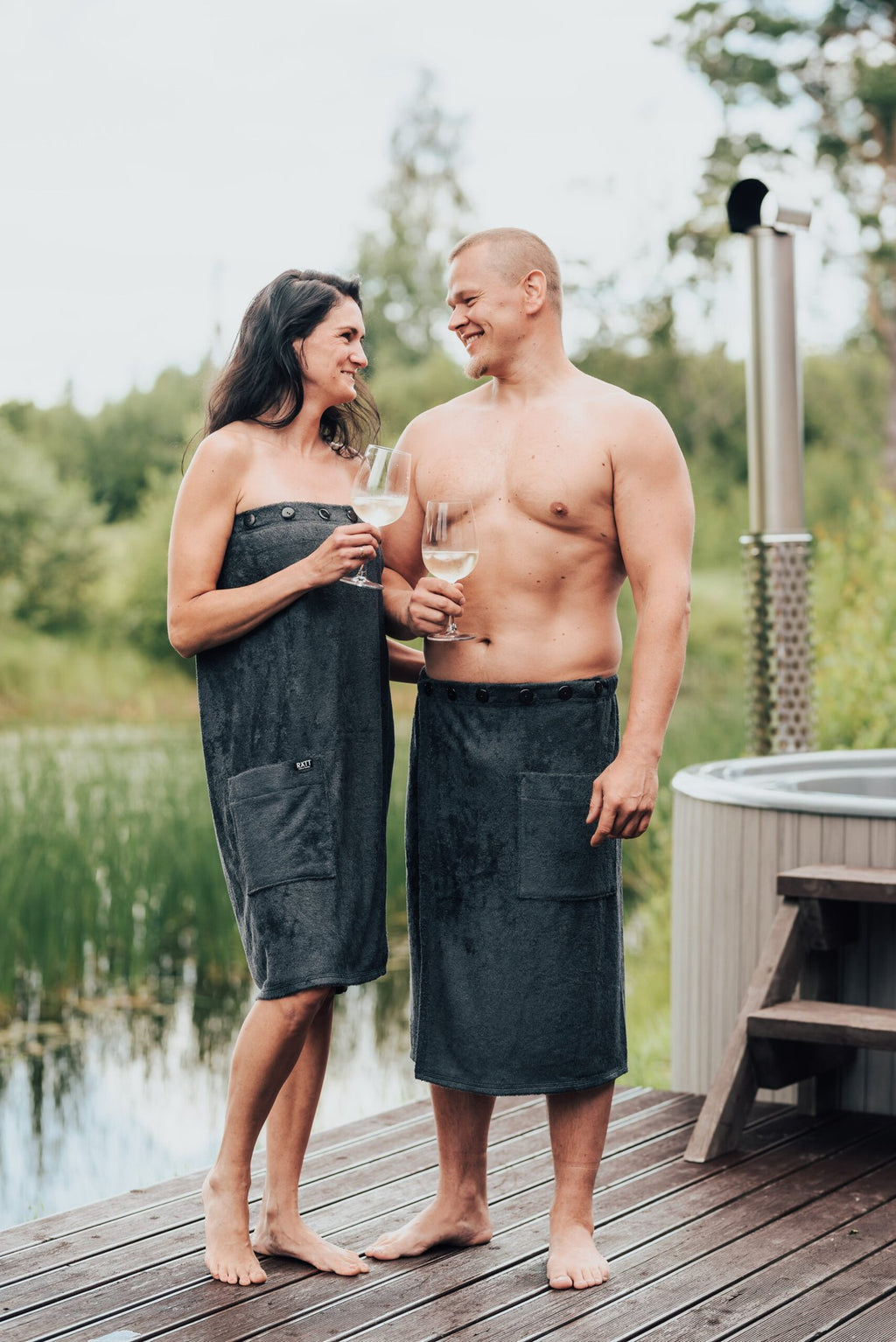 1. Women's dark grey bamboo terry spa wrap by RÄTT, worn by female and male models by a hot tub, showcasing adjustable fit and pocket
