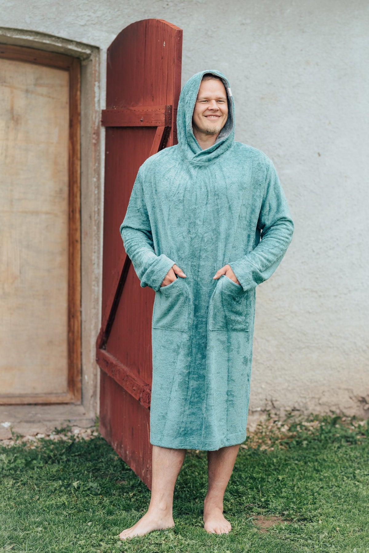 8. Men's green bamboo terry long-sleeve poncho with hood, worn by model standing outdoors near a wooden door
