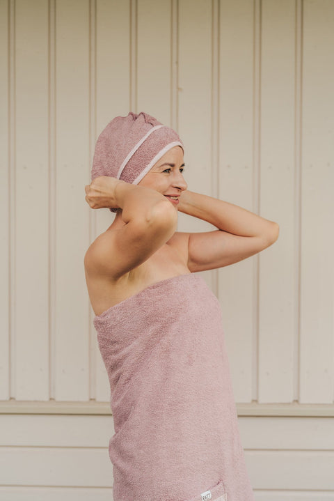 1. Woman wearing RÄTT cotton terry hair wrap in pink, adjusting it while wrapped in matching towel, lifestyle setting