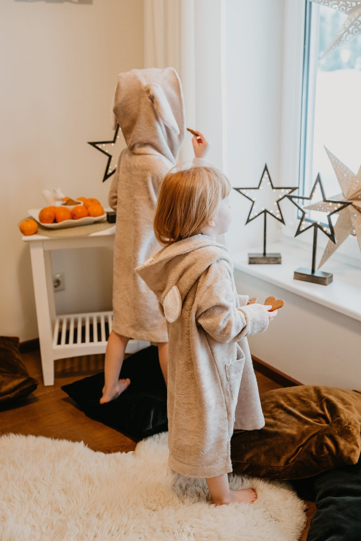 2. Two children in beige RÄTT Long-Sleeve Poncho TEDDY with hoods, standing by a window with star decorations