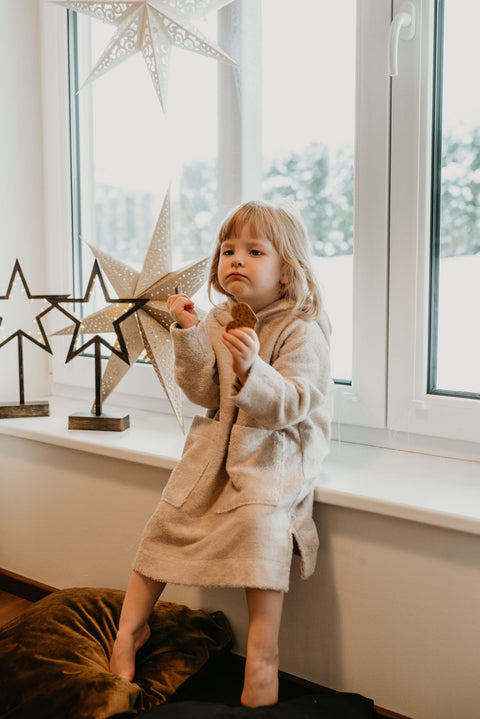 5. Toddler sitting on a windowsill wearing RÄTT long-sleeve poncho TEDDY in beige, holding a cookie