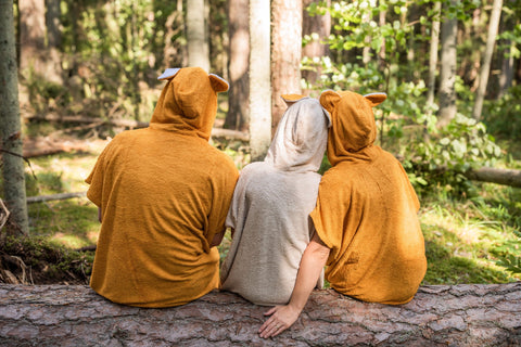3. Back view of unisex bamboo terry ponchos in orange and beige worn by three models sitting on a log