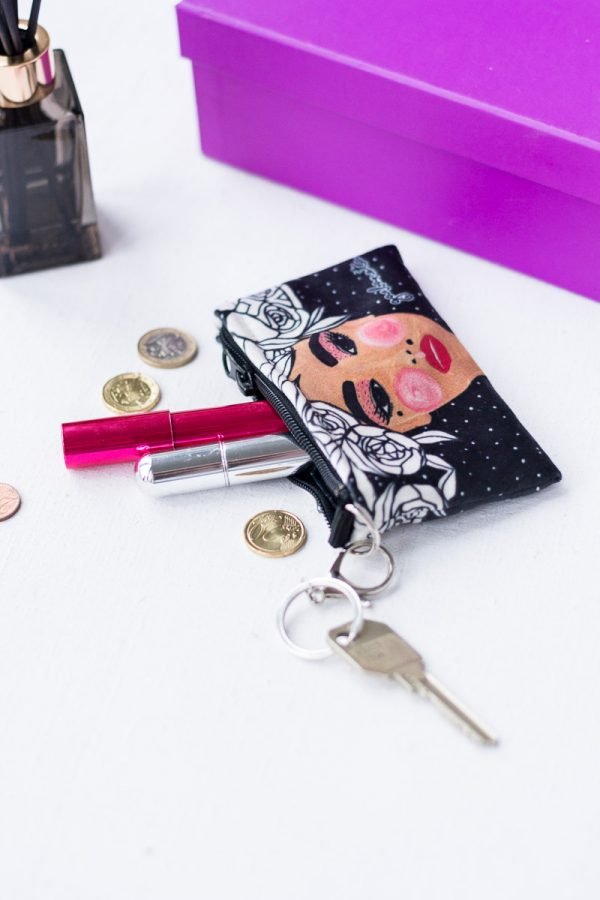 5. Garbanota Key Pouch Yoko in black with artistic face design, shown with keyring, coins, and lipstick on a white surface