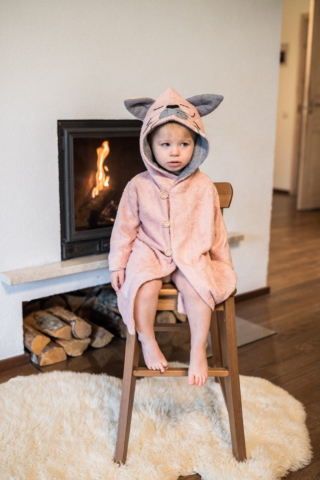 1. Toddler wearing pink RÄTT Bamboo Terry Bathrobe CAT with hood, sitting by fireplace