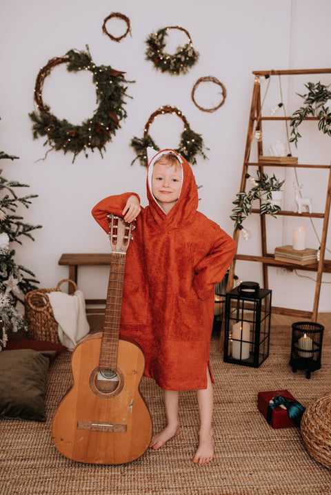 2. Toddler in RÄTT Long-Sleeve Poncho FOX with hood, standing next to guitar in festive setting
