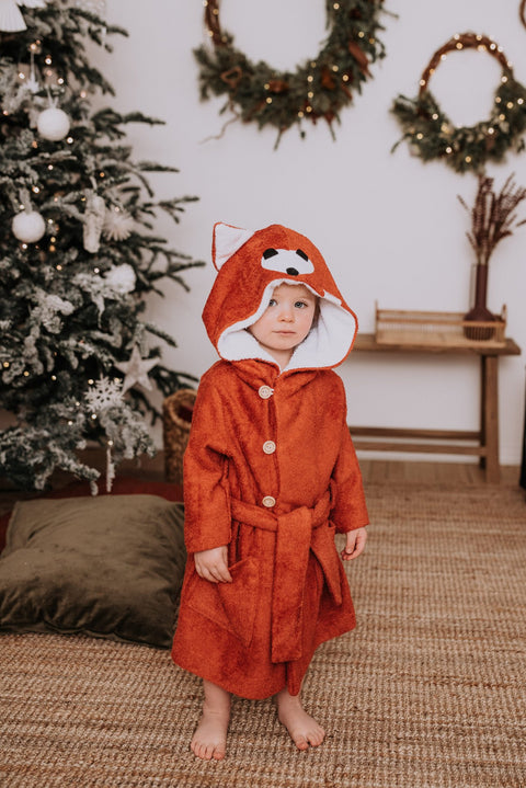 1. Toddler wearing RÄTT FOX Bamboo Terry Bathrobe with hood and button closure, standing in a cozy room with holiday decor
