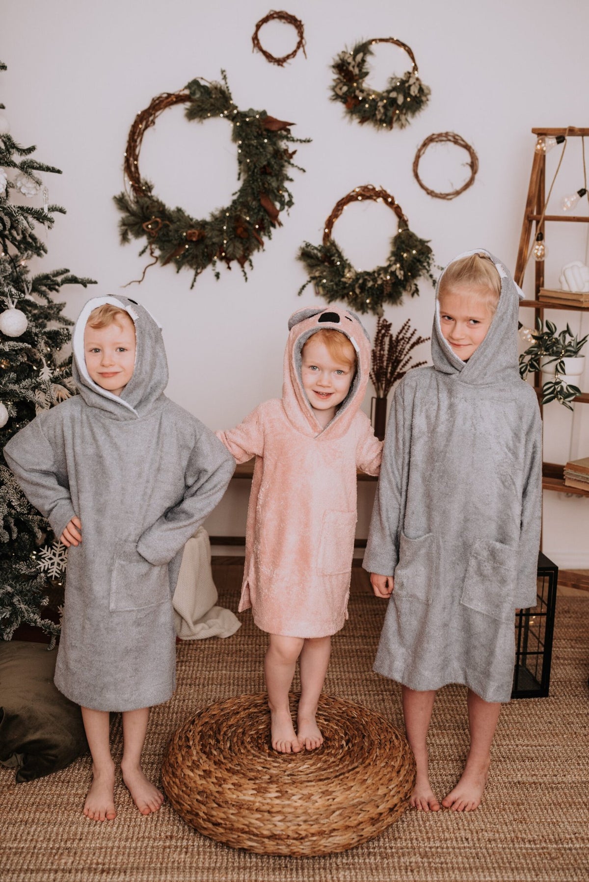 1. Three children wearing RÄTT long-sleeve ponchos in grey and pink, standing indoors with holiday decor