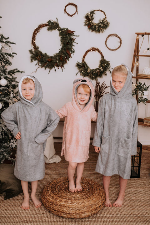 1. Three children wearing RÄTT long-sleeve ponchos in grey and pink, standing indoors with holiday decor