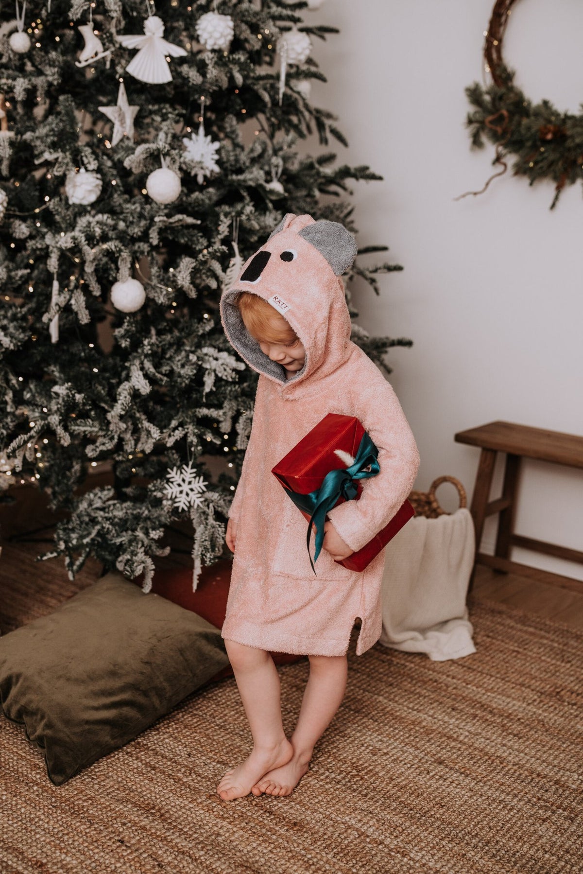 2. Child in pink RÄTT long-sleeve poncho with koala hood holding a gift, standing by a decorated Christmas tree