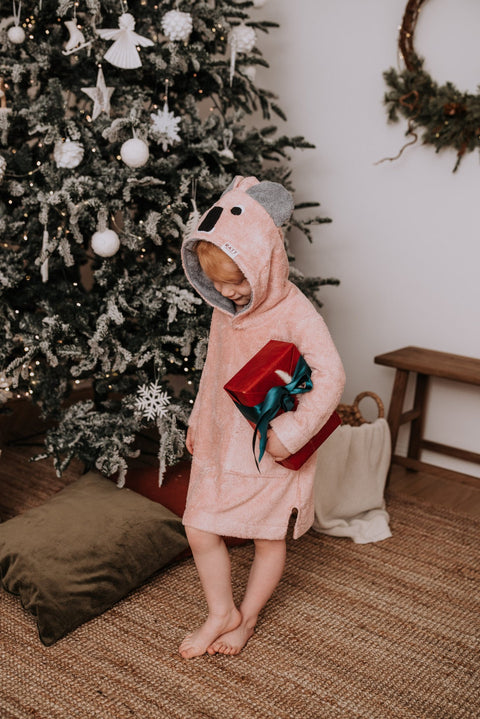 2. Child in pink RÄTT long-sleeve poncho with koala hood holding a gift, standing by a decorated Christmas tree