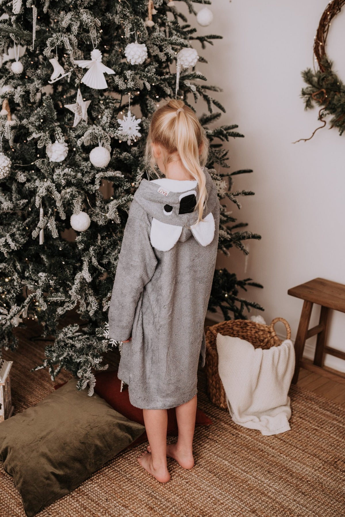 3. Child wearing grey RÄTT long-sleeve poncho with koala hood, shown from the back near a Christmas tree