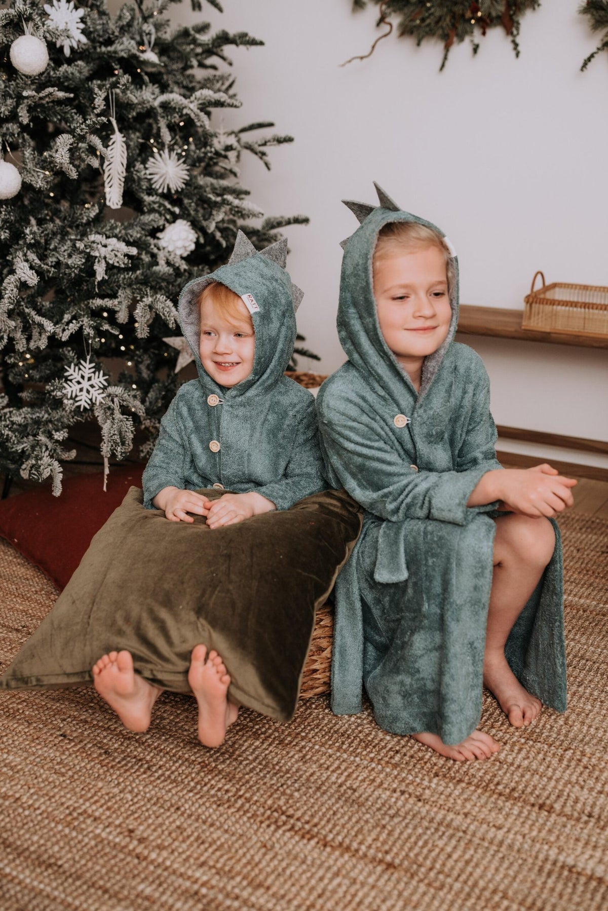 1. Two toddlers wearing RÄTT's dinosaur-themed bamboo terry bathrobes, sitting by a Christmas tree, showcasing cozy hood and button closure