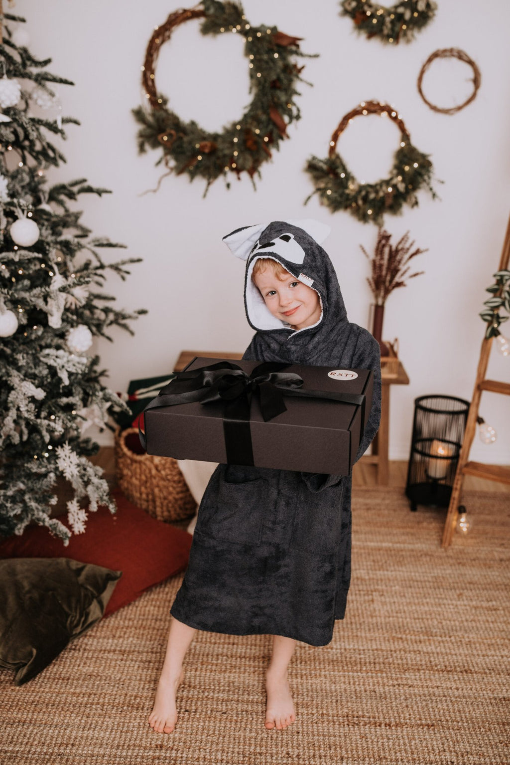 1. Toddler wearing RÄTT Long-Sleeve Poncho WOLF in cozy indoor setting, holding a gift box
