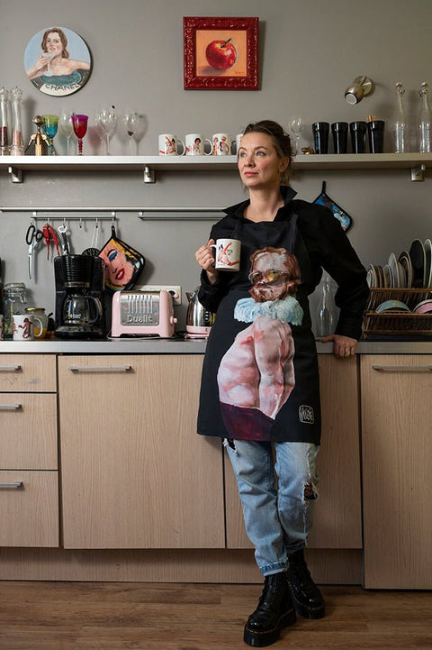 1. Woman in kitchen wearing Katrin Valgemäe's 'Pervert in the Kitchen' apron, holding a mug, surrounded by eclectic decor