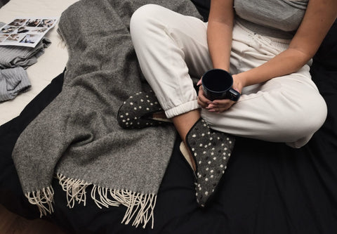 1. Woman wearing Omaking Barefoot Wool Slippers Trolla with dots, sitting on bed with blanket and holding a mug