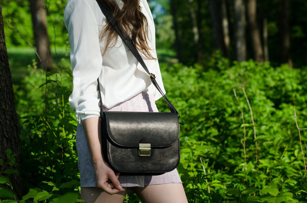 1. Woman holding Zelma Kraft black leather saddle bag with adjustable strap, shown in lifestyle setting