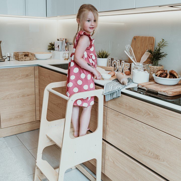 17. Child in pink dress on natural wood Montessori helper tower step stool in kitchen