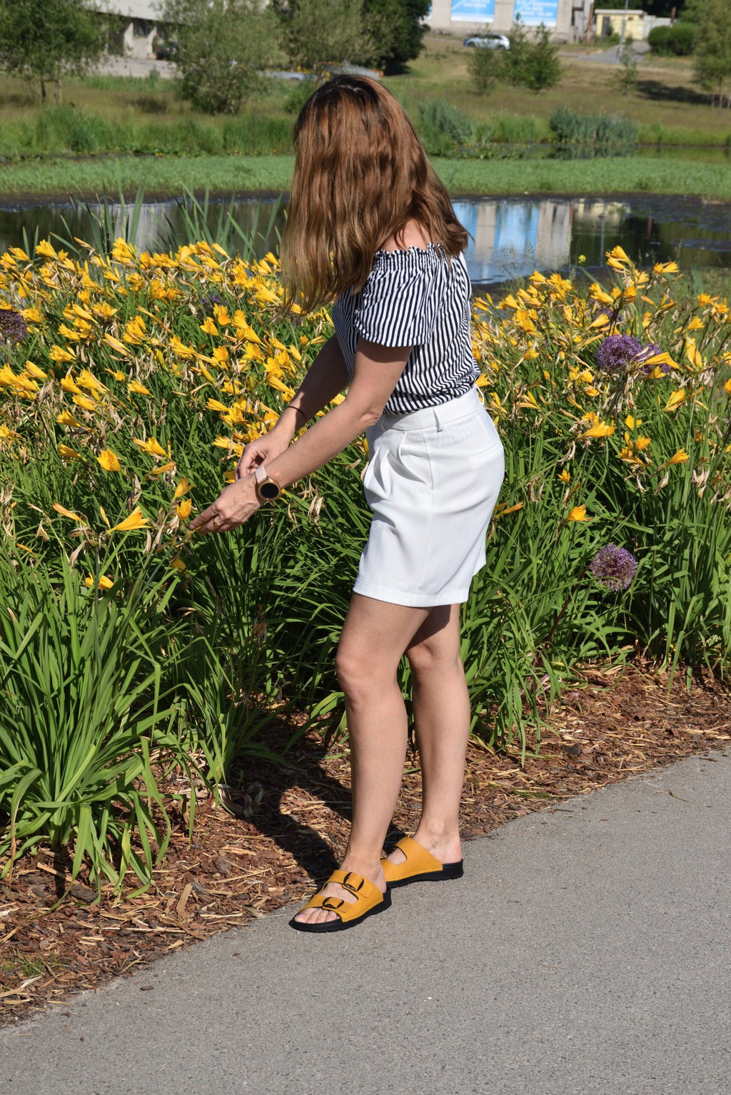 1. Woman wearing Meeliku Ocre Sandals by Omaking, styled with white shorts and striped top, outdoors near flowers