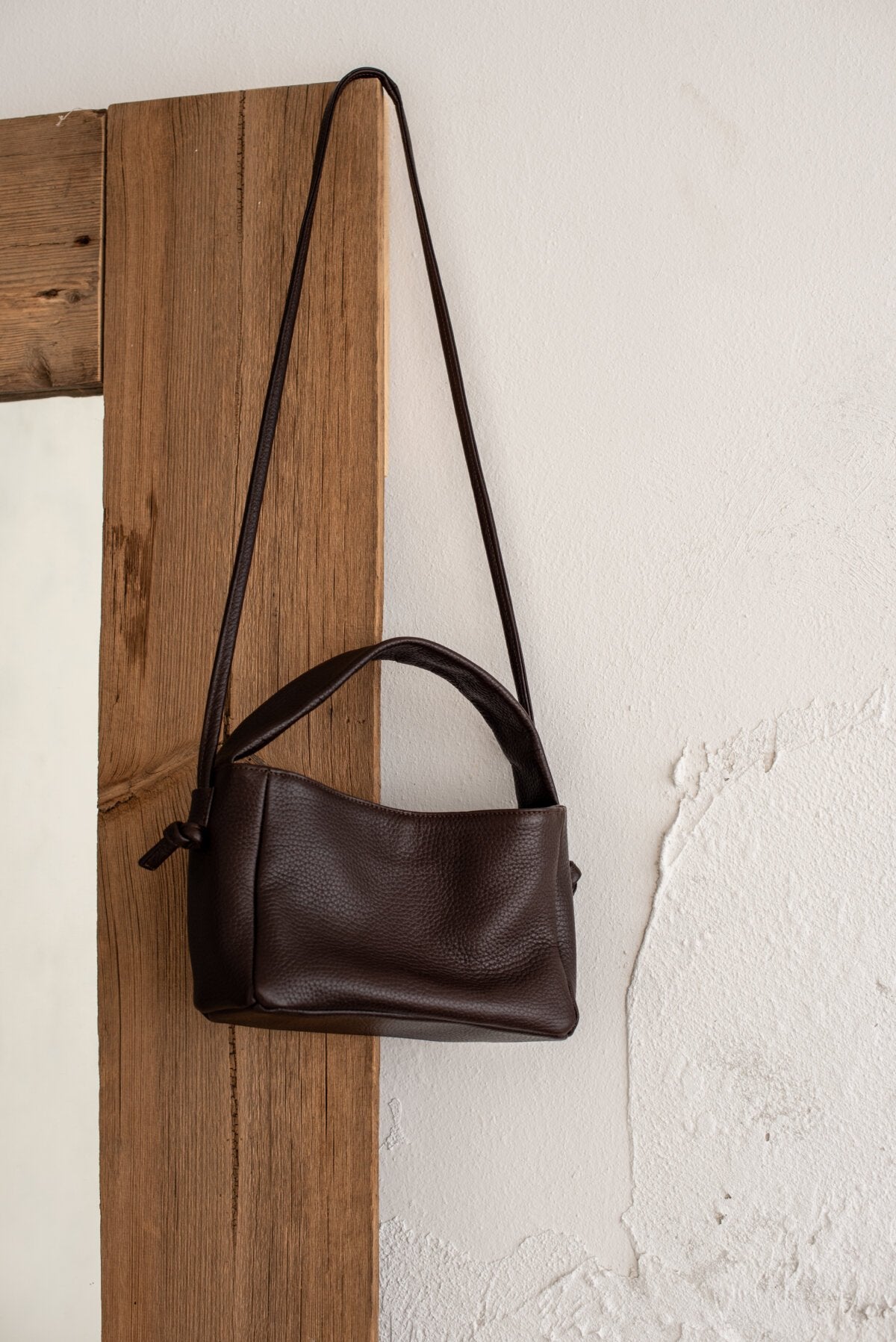 6. Lava flow Crossbody Bag MERU in dark brown leather hanging on a wooden door