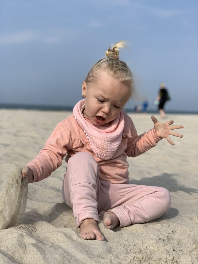 1. Child wearing light pink muslin scarf-bib and matching outfit playing on sandy beach