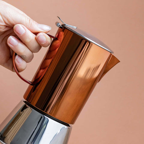 1. Hand holding La Cafetière copper and silver stainless steel stovetop coffee maker against a peach background