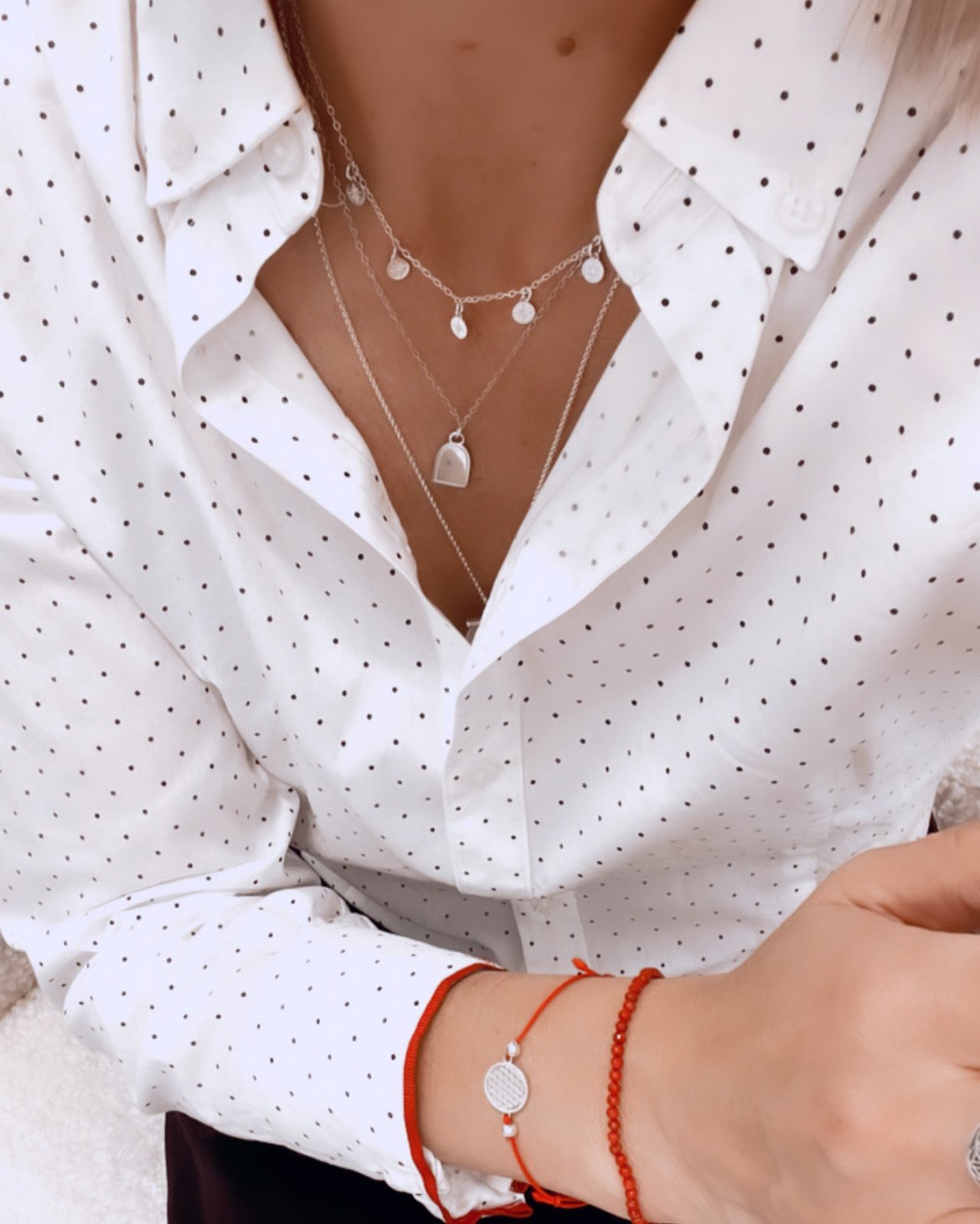 1. Woman wearing New Vintage by Kriss Flower Of Life Bracelet with red cord and silver charm, styled with layered necklaces and polka dot shirt
