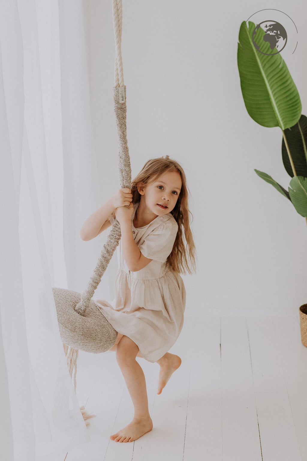 2. Young girl in beige dress swinging on Nofi KIDS bouclé sand swing indoors with green plant in background