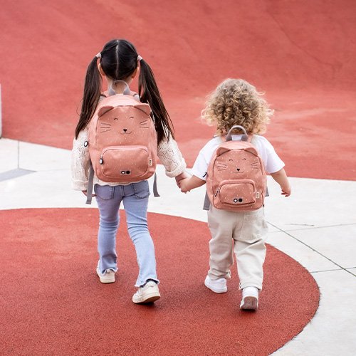 1. Two children wearing Trixie Baby Mrs Cat backpacks, walking hand in hand on a playground