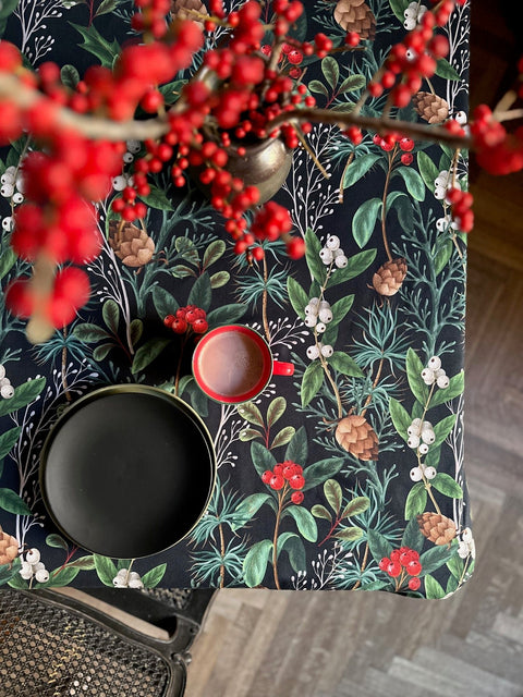 1. Festive Motive Christmas Forest tablecloth by Hortensias Home, featuring a vibrant holiday design with red berries and pinecones, styled with a red mug and black plate