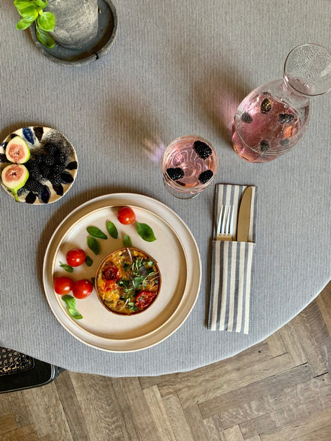 1. Lifestyle image of Nord 2 round tablecloth by Hortensias Home with subtle blue lines on beige, set with a meal and drinks