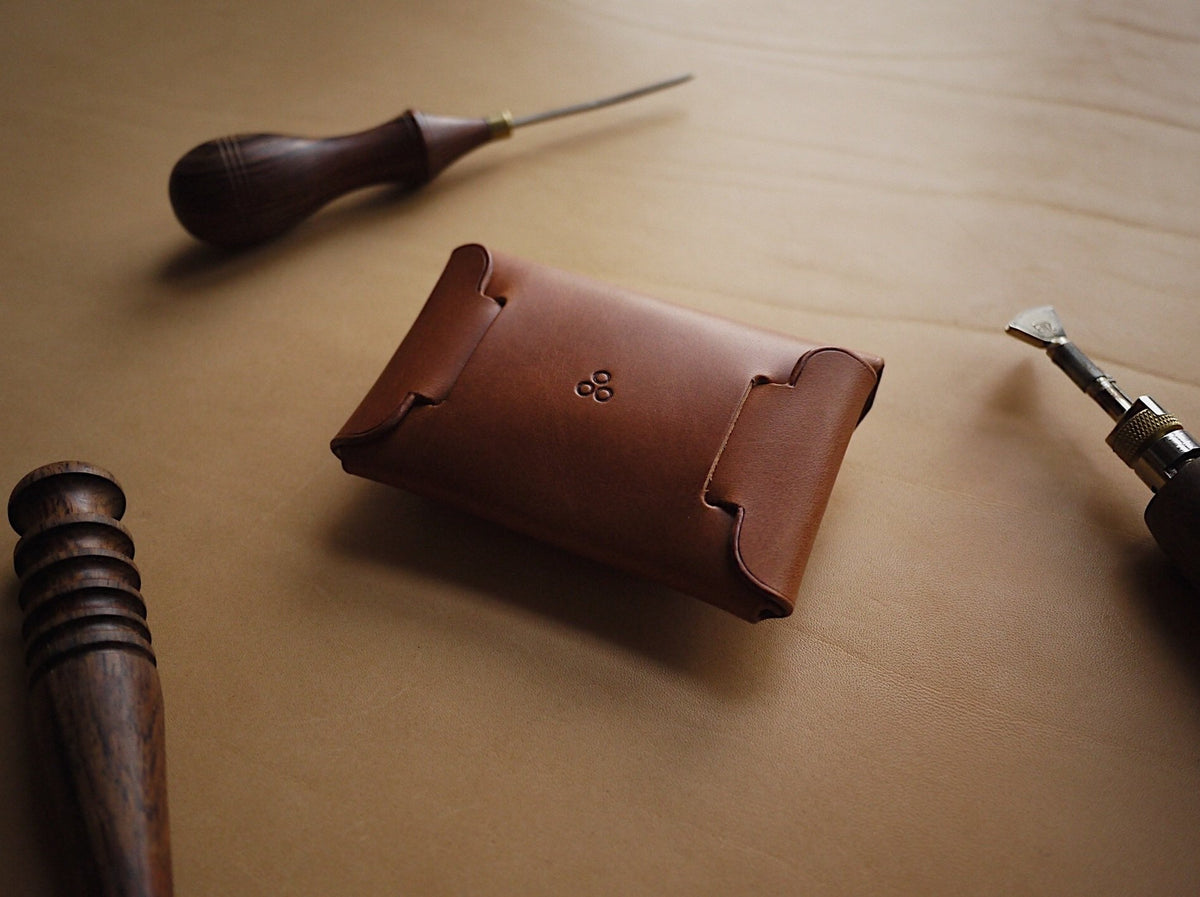 1. Mirgoods Button Leather Wallet in cognac, shown closed with polished edges and creasing lines, surrounded by leather crafting tools