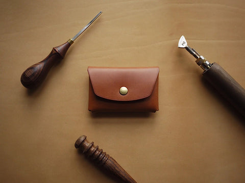 2. Mirgoods Button Leather Wallet in cognac, front view with brass snap button, displayed with leather crafting tools