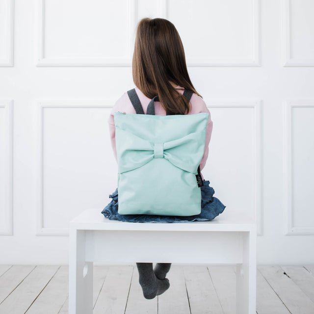 1. Girl wearing Muni kids waterproof mint bow backpack, sitting on a white bench in a lifestyle setting