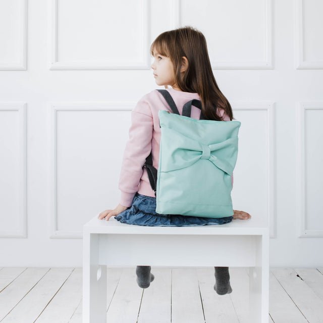 2. Girl with Muni kids mint bow backpack, sitting sideways on a white bench in a lifestyle setting