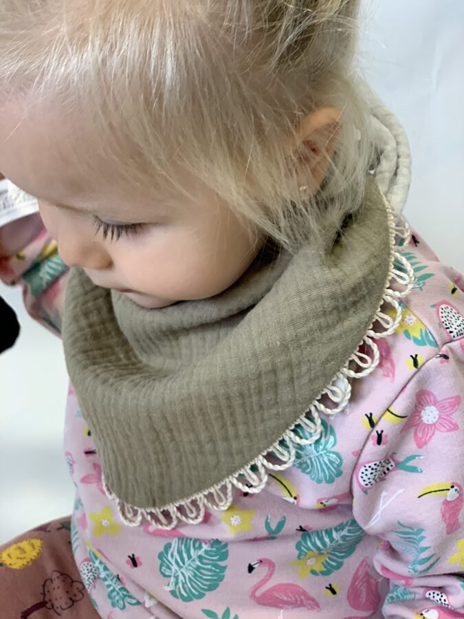 1. Child wearing khaki muslin scarf-bib with lace trim, paired with a colorful floral outfit, sitting indoors